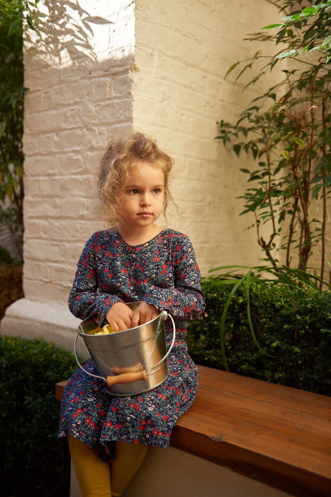 A girl sitting on a bench in the nursery garden with a metal bucket