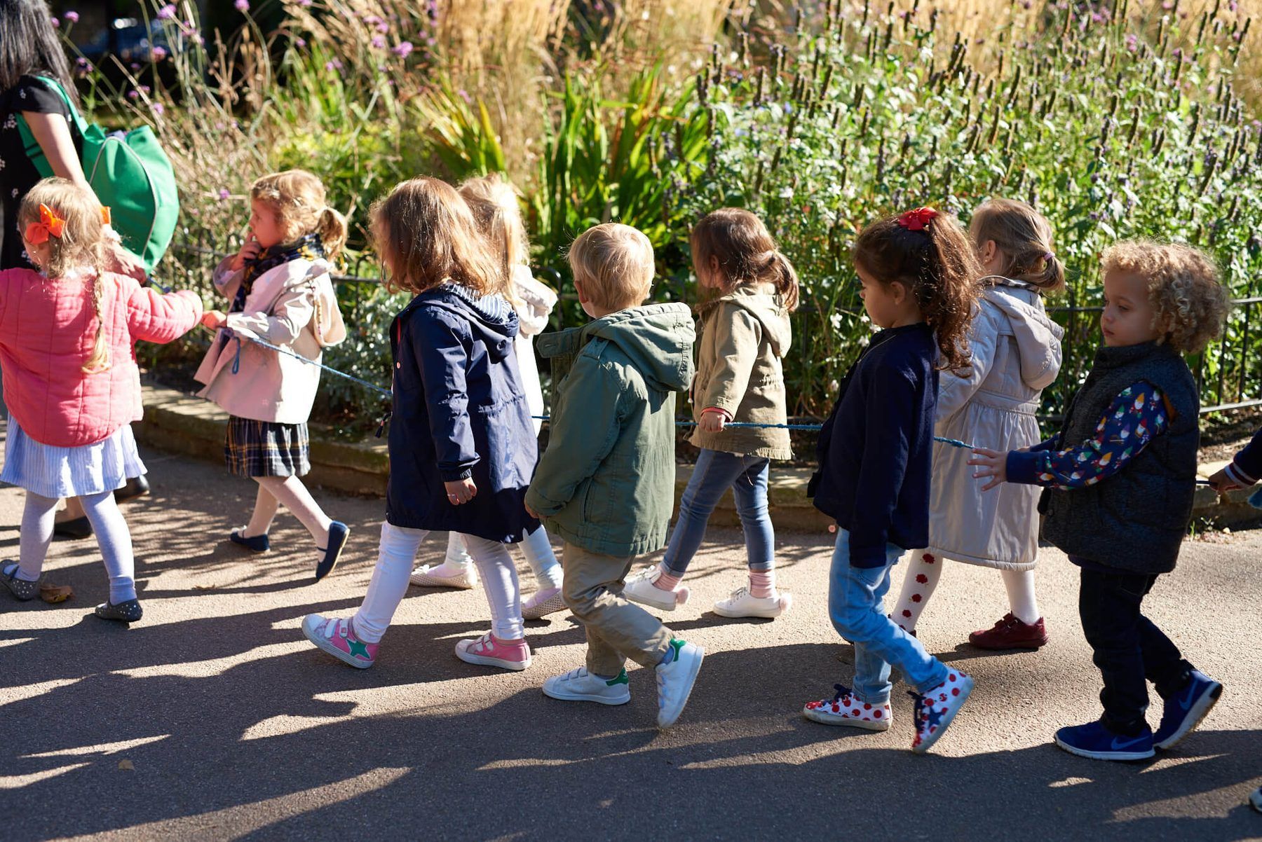 A line of children in coats walking through a sunny garden square